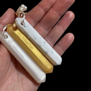 May include: Four rectangular pendants in white, gold, and speckled white, each with a small loop and cord. The pendants are engraved with the text "Willows Toys". The pendants are held in a hand against a black background.