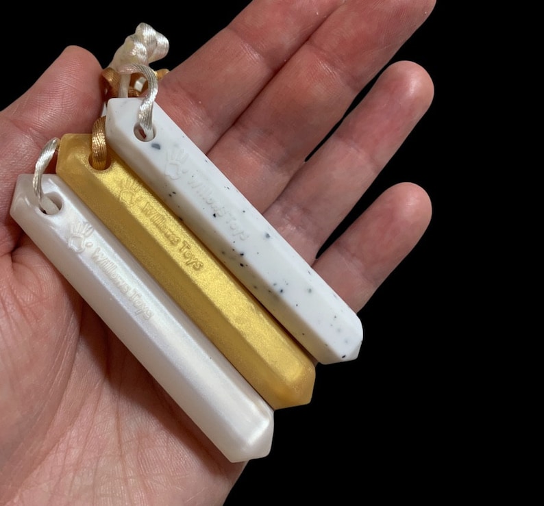 May include: Four rectangular pendants in white, gold, and speckled white, each with a small loop and cord. The pendants are engraved with the text "Willows Toys". The pendants are held in a hand against a black background.
