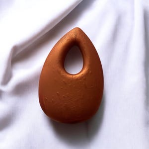 May include: A teardrop-shaped pendant with a hole in the center. The pendant is a metallic copper color and has a textured surface. The pendant is displayed on a white background.