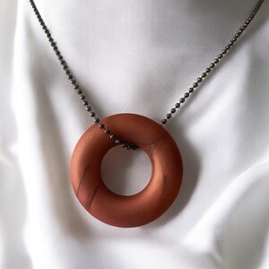 May include: A copper-colored, circular pendant necklace. The pendant is a solid ring shape, suspended from a delicate, dark metal ball chain. The background is a white, silky fabric.
