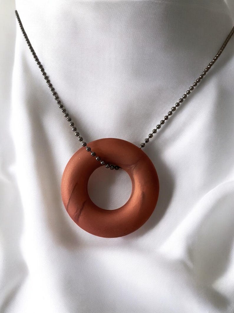 May include: A copper-colored, circular pendant necklace. The pendant is a solid ring shape, suspended from a delicate, dark metal ball chain. The background is a white, silky fabric.