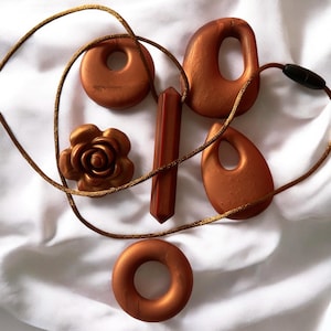 May include: A collection of bronze-colored jewelry pieces, including a rose, a donut-shaped pendant, and various abstract shapes. A bronze cord weaves through the pieces, suggesting necklaces. The items are arranged on a white fabric background.
