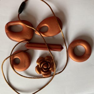 May include: A collection of copper-colored pendants and beads, including a flower, a donut shape, and a geometric bar. The pendants are strung on a brown cord with a black clasp. The jewelry pieces are arranged on a white surface.