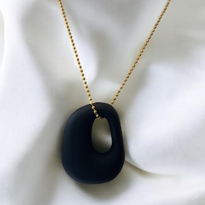 Puede incluir: Un collar de cadena dorada con un colgante ovalado negro liso que presenta un recorte. El colgante tiene una forma irregular con un agujero en el centro.