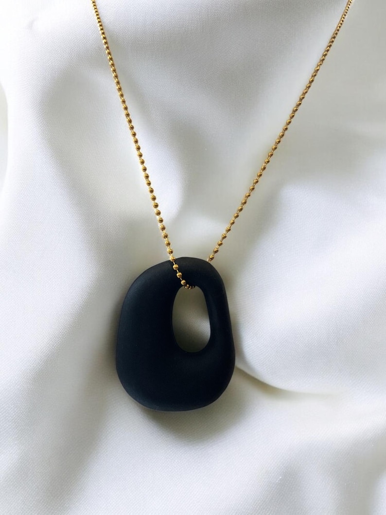Puede incluir: Un collar de cadena dorada con un colgante ovalado negro liso que presenta un recorte. El colgante tiene una forma irregular con un agujero en el centro.