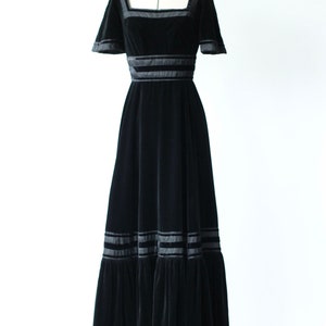 May include: A black velvet maxi dress with a square neckline and short sleeves. The dress has a fitted bodice and a flowing skirt with multiple layers of ruffles. The dress is accented with black satin ribbon trim.
