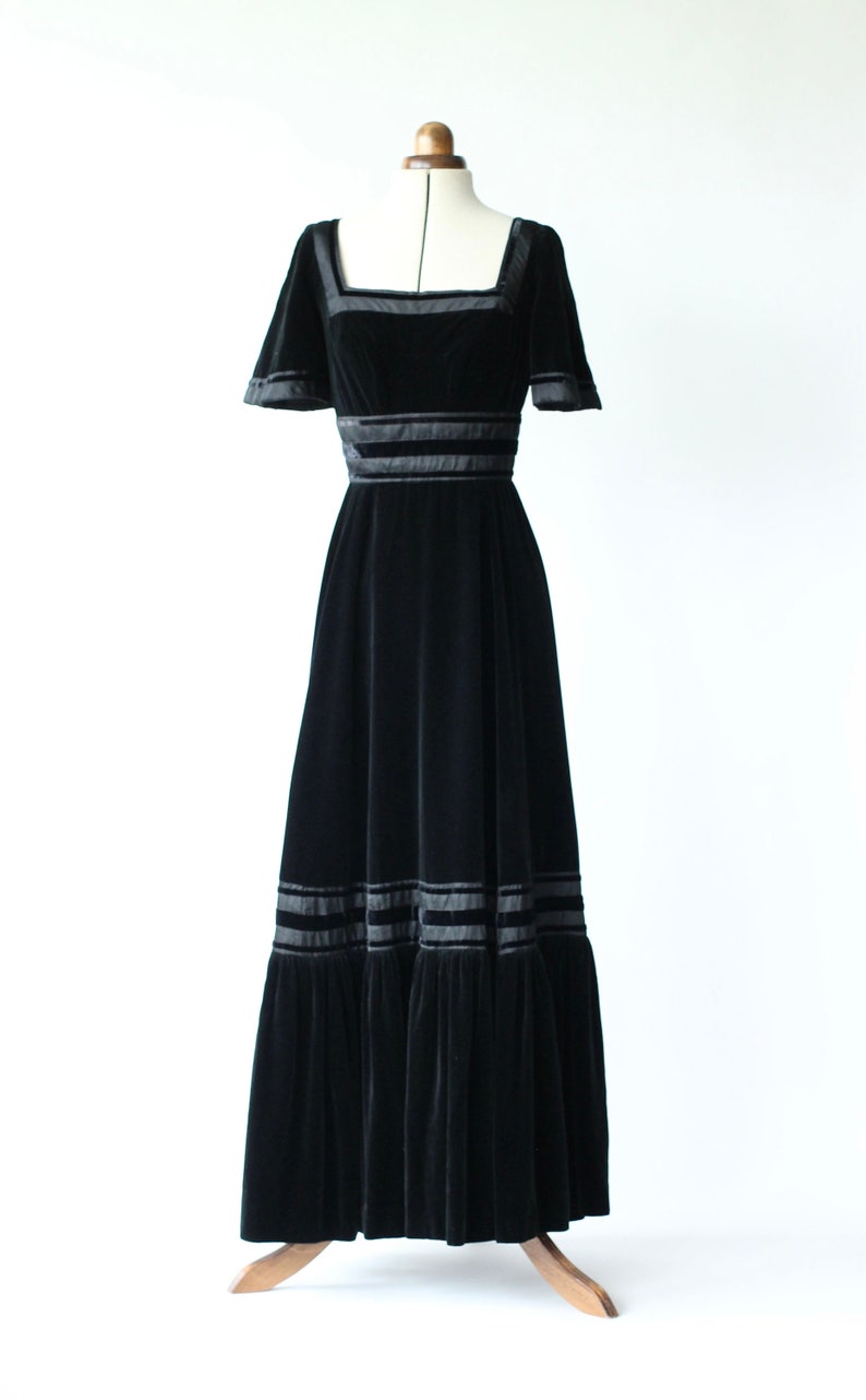 May include: A black velvet maxi dress with a square neckline and short sleeves. The dress has a fitted bodice and a flowing skirt with multiple layers of ruffles. The dress is accented with black satin ribbon trim.