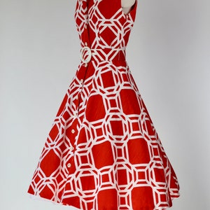 May include: A red sleeveless dress with a white geometric pattern and a white belt. The dress has a collared neckline and a button-up front.
