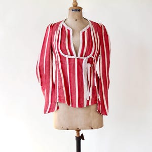 May include: A red and white striped blouse with long sleeves and a tie closure. The blouse is displayed on a vintage dress form. The vertical stripes are evenly spaced and run the length of the garment. The neckline is a V-shape.
