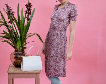 Könnte beinhalten: Ein Vintage-Kleid mit floralem Muster in Rosa- und Lilatönen mit kurzen Ärmeln und V-Ausschnitt. Eine weiße Handtasche mit goldenem Verschluss steht auf einem kleinen Holzschemel. Der Hintergrund ist einfarbig rosa. Die Person trägt blaue Strumpfhosen.