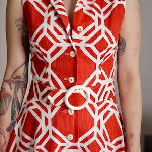 May include: A sleeveless red and white geometric print dress with a white belt and buttons.