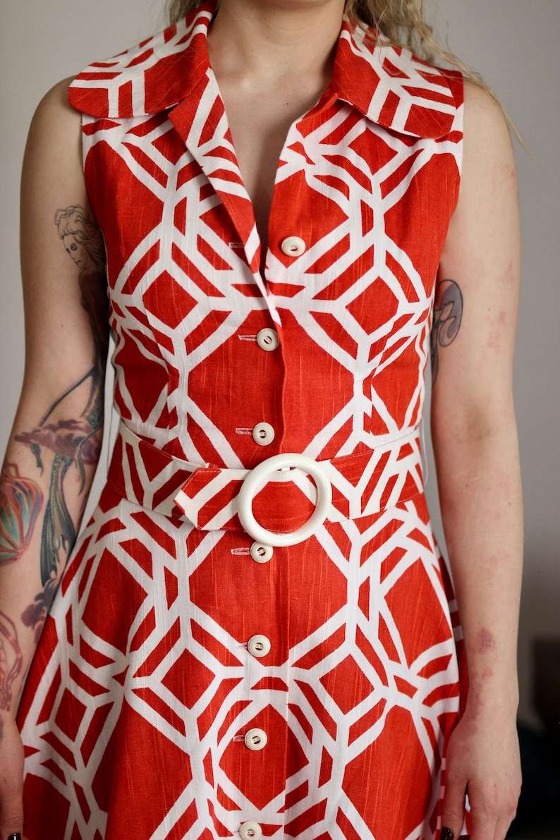 May include: A sleeveless red and white geometric print dress with a white belt and buttons.