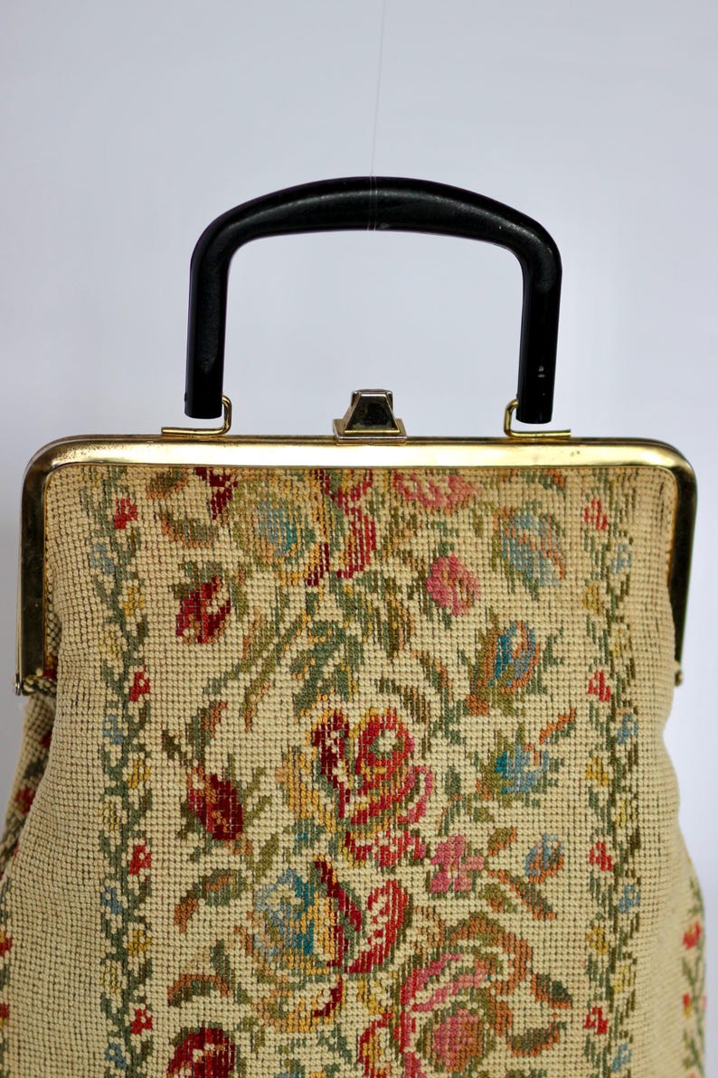 May include: Vintage handbag with a black handle and gold-toned frame. The beige fabric is intricately embroidered with a floral pattern in shades of red, pink, blue, and green. The bag has a classic, structured design.