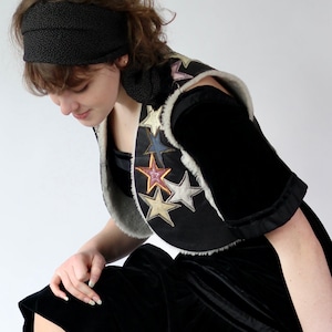 May include: Black velvet vest with colorful star appliqués and a faux fur trim. The vest is paired with a black headband and a black velvet dress. The stars are in gold, silver, pink, and blue.