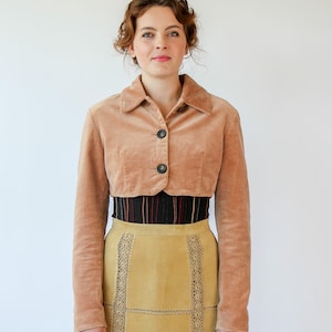 May include: A light brown cropped jacket with two dark buttons, worn with a high-waisted, knee-length yellow skirt. The skirt has decorative stitching and a black and red striped band at the waist. The outfit is set against a plain white background.