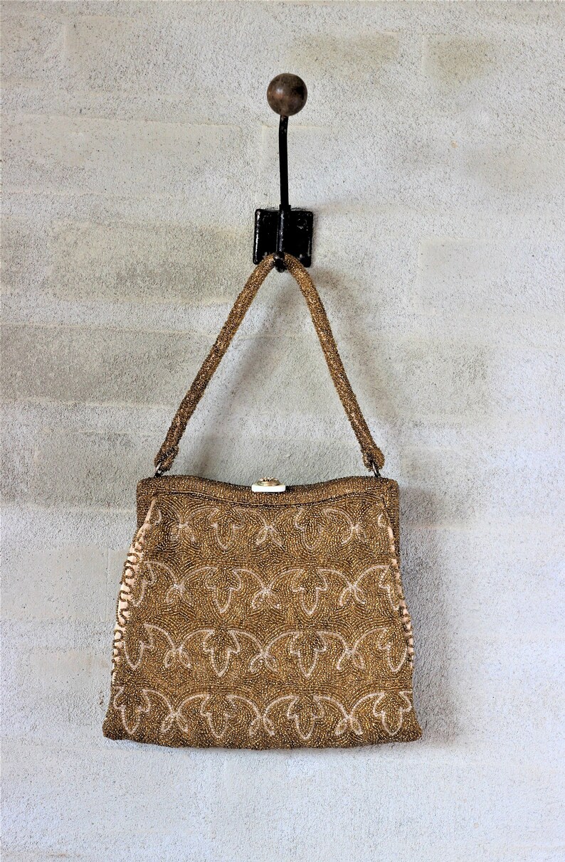 Puede incluir: Un bolso de mano de cuentas doradas con un dise&ntilde;o floral cuelga de un gancho negro contra una pared blanca.