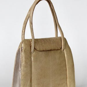 May include: A beige handbag with a reptile-effect texture. The bag features a rectangular design, a flap closure, and two rounded handles. The interior lining is a lighter beige colour.