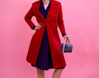 May include: A woman wearing a red coat over a purple dress, paired with red shoes and earrings. She holds a small, dark blue handbag. The outfit is set against a pink backdrop, creating a vintage aesthetic.