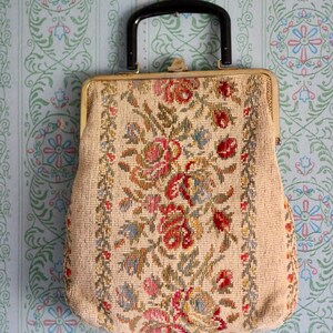 May include: Vintage beige handbag with a black handle and gold-tone clasp. The bag features a floral needlepoint design in red, pink, blue, and green. The background is a light blue wallpaper with a floral pattern.