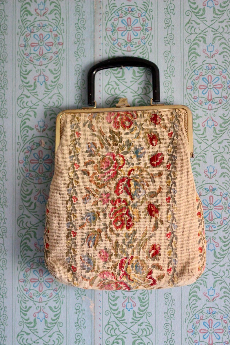 May include: Vintage beige handbag with a black handle and gold-tone clasp. The bag features a floral needlepoint design in red, pink, blue, and green. The background is a light blue wallpaper with a floral pattern.