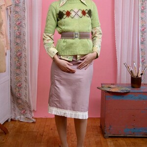 Puede incluir: Una mujer con un atuendo vintage. El atuendo incluye un suéter verde claro con un patrón de diamantes, una falda rosa y zapatos verdes. El fondo es una pared rosa con cortinas florales.