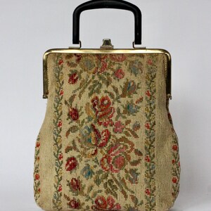 May include: Vintage tapestry handbag with a black handle and gold-toned frame. The beige fabric is adorned with a floral pattern in red, pink, green, and blue. The bag has a structured shape and a clasp closure.
