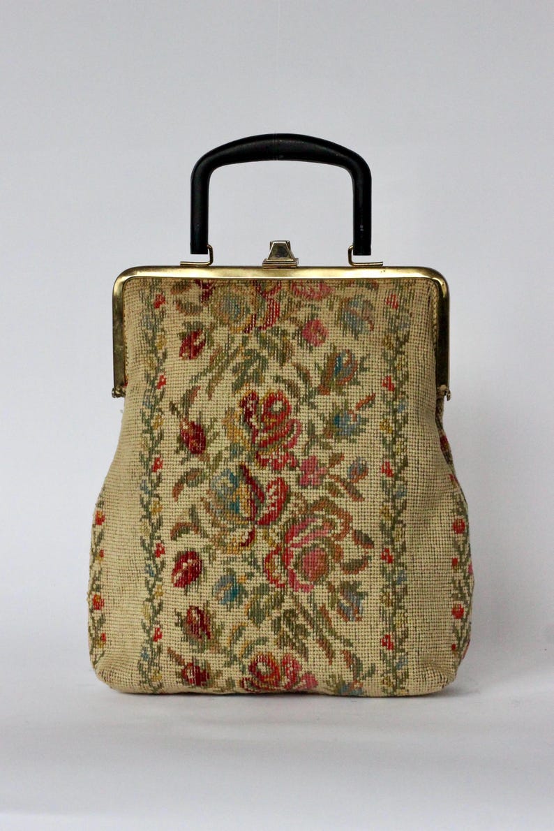 May include: Vintage tapestry handbag with a black handle and gold-toned frame. The beige fabric is adorned with a floral pattern in red, pink, green, and blue. The bag has a structured shape and a clasp closure.