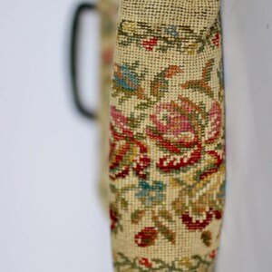 May include: A vintage-style handbag with a floral tapestry design. The bag features a cream-colored background with a detailed pattern of red, pink, blue, and green flowers and leaves. A black handle is visible on the side.