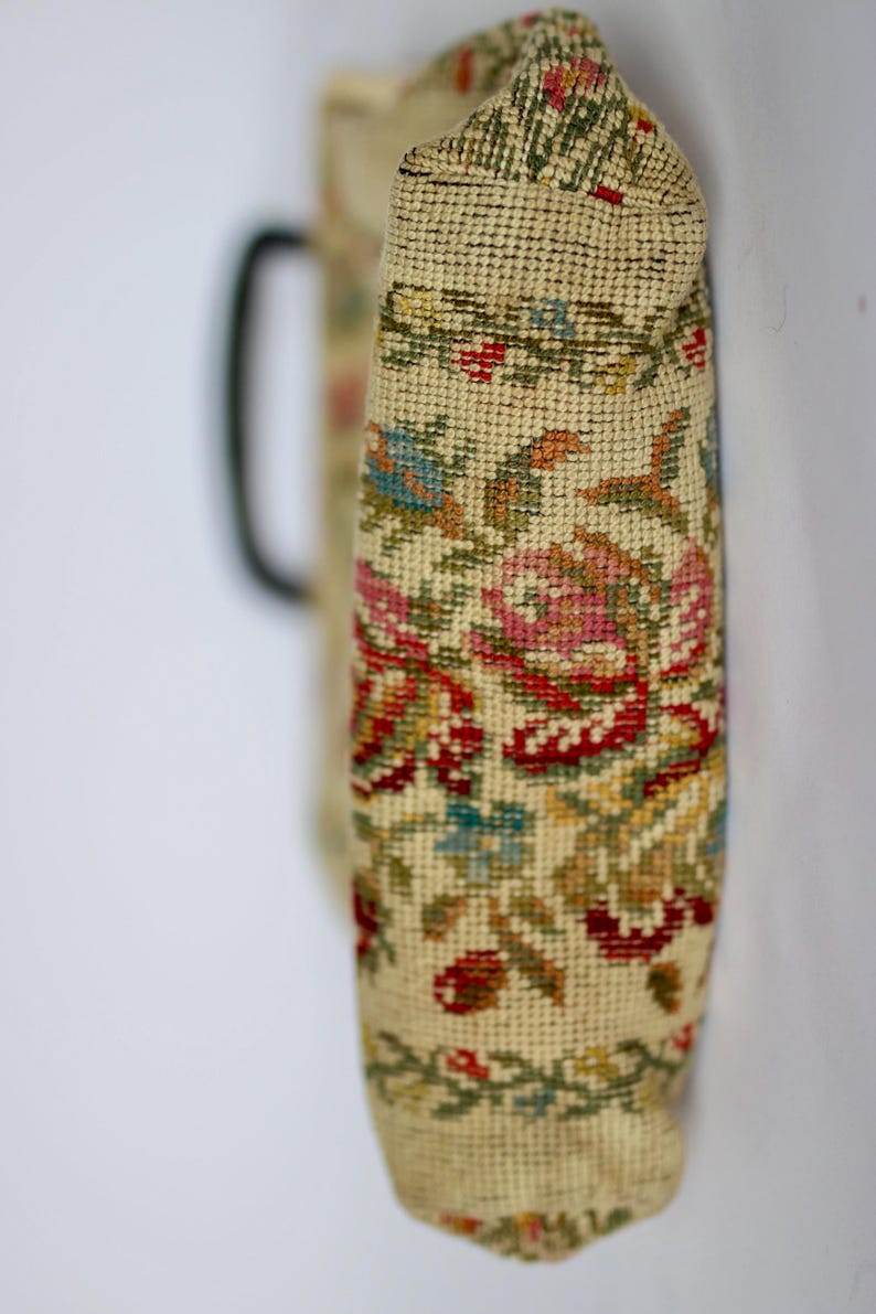 May include: A vintage-style handbag with a floral tapestry design. The bag features a cream-colored background with a detailed pattern of red, pink, blue, and green flowers and leaves. A black handle is visible on the side.
