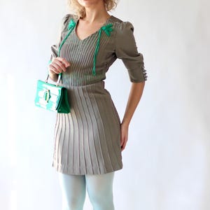 May include: A vintage-style outfit featuring a gray pleated dress with a V-neck and short sleeves. The dress is paired with light blue tights and a bright green handbag. Sunglasses rest on top of the head.