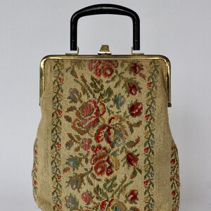 May include: Vintage tapestry handbag with a black handle and gold-toned clasp. The beige fabric is adorned with a floral pattern in red, pink, blue, and green. The bag has a rectangular shape with a structured design, perfect for carrying essentials.