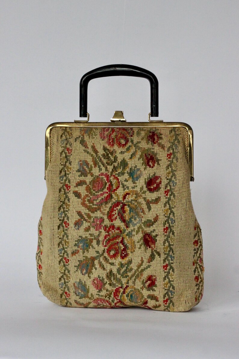 May include: Vintage tapestry handbag with a black handle and gold-toned clasp. The beige fabric is adorned with a floral pattern in red, pink, blue, and green. The bag has a rectangular shape with a structured design, perfect for carrying essentials.