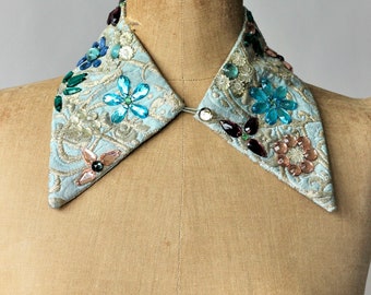 Puede incluir: Un cuello de tela azul claro con forma puntiaguda y adornado con brillantes y lentejuelas de colores.