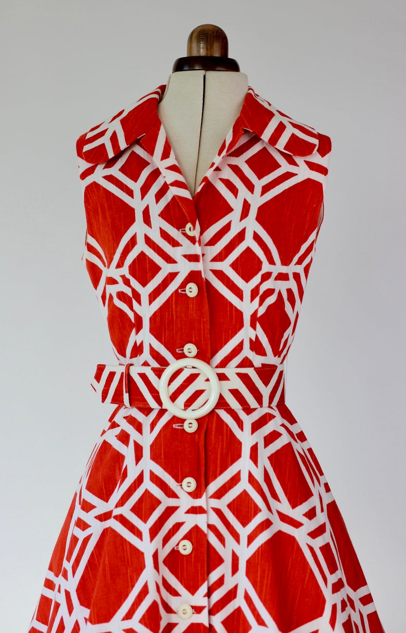 May include: A sleeveless orange and white geometric print dress with a white belt and buttons down the front.