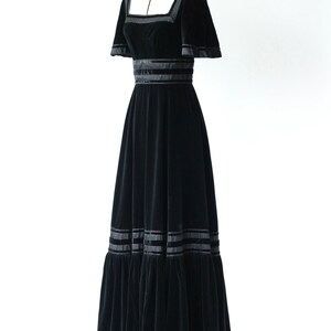 May include: A black velvet maxi dress with a square neckline and short sleeves. The dress has a fitted bodice and a flowing skirt with multiple layers of ruffles. The dress is decorated with black satin ribbon trim.