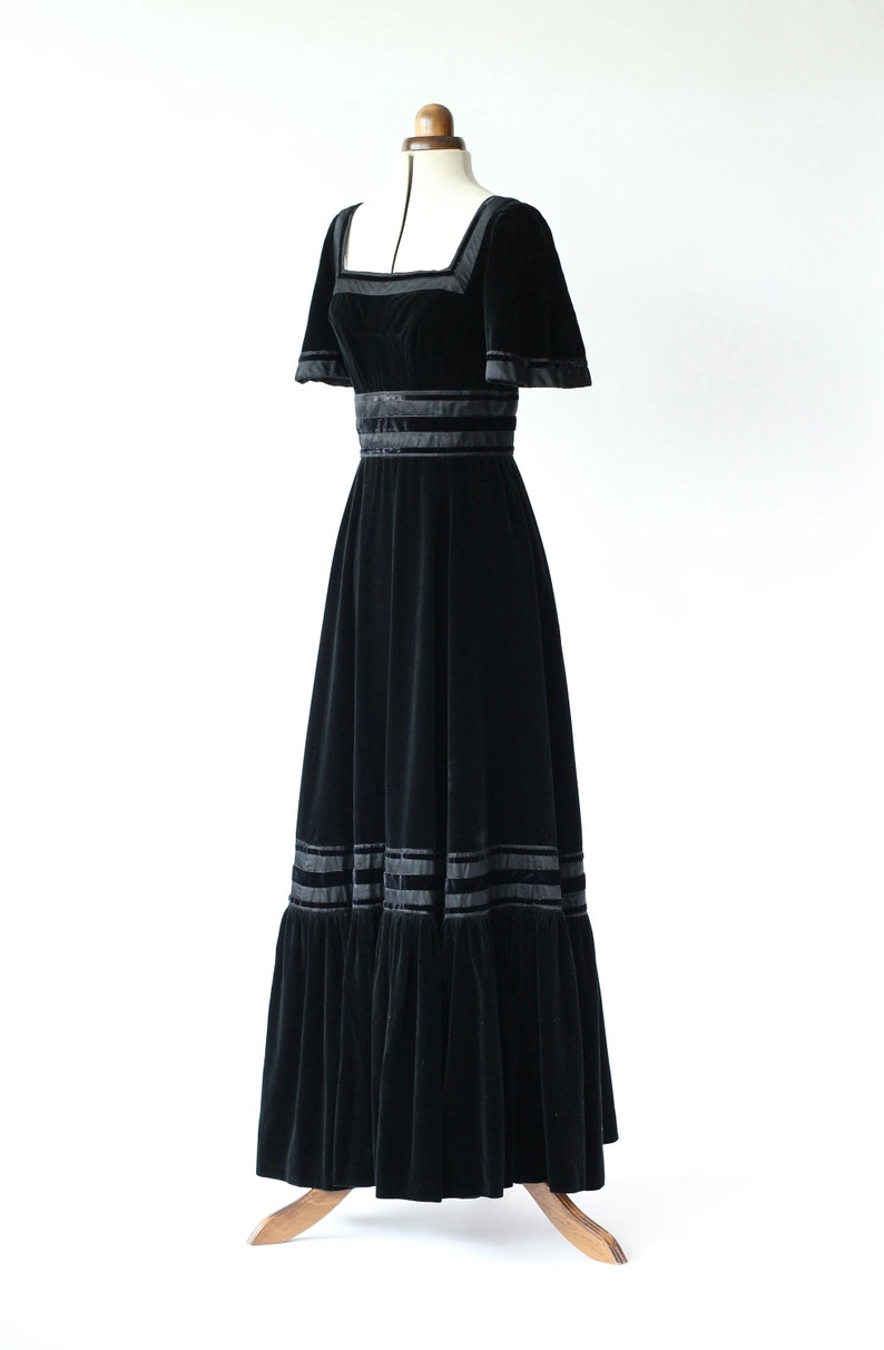 May include: A black velvet maxi dress with a square neckline and short sleeves. The dress has a fitted bodice and a flowing skirt with multiple layers of ruffles. The dress is decorated with black satin ribbon trim.