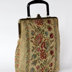 May include: Vintage beige handbag with a black handle and gold-toned clasp. The bag features a floral tapestry design in shades of red, pink, green, and blue. The sides have a vertical floral border. A classic, elegant accessory.