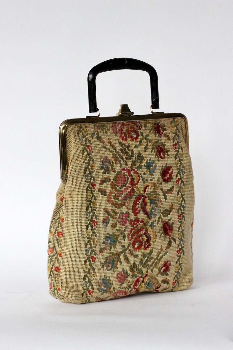 May include: Vintage beige handbag with a black handle and gold-toned clasp. The bag features a floral tapestry design in shades of red, pink, green, and blue. The sides have a vertical floral border. A classic, elegant accessory.
