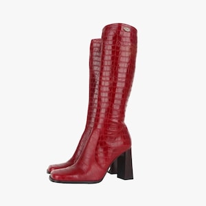 Puede incluir: Botas rojas hasta la rodilla con textura de piel de cocodrilo sintética. Las botas tienen punta cuadrada y tacón grueso marrón oscuro. Un pequeño emblema plateado es visible en la parte superior de la bota.