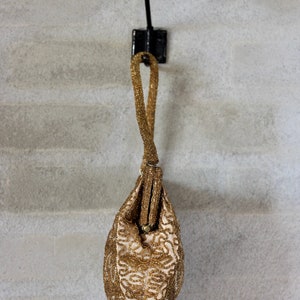 Puede incluir: Un bolso de noche de cuentas doradas cuelga de un gancho negro en una pared blanca. El bolso tiene un dise&ntilde;o delicado e intrincado y una larga correa de cuentas.