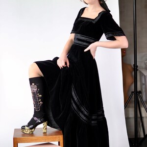 May include: A woman wearing a black velvet dress with a black velvet headband. She is standing on a wooden stool with two boxes stacked on top of each other. She is wearing black socks with gold embroidery and black platform shoes.