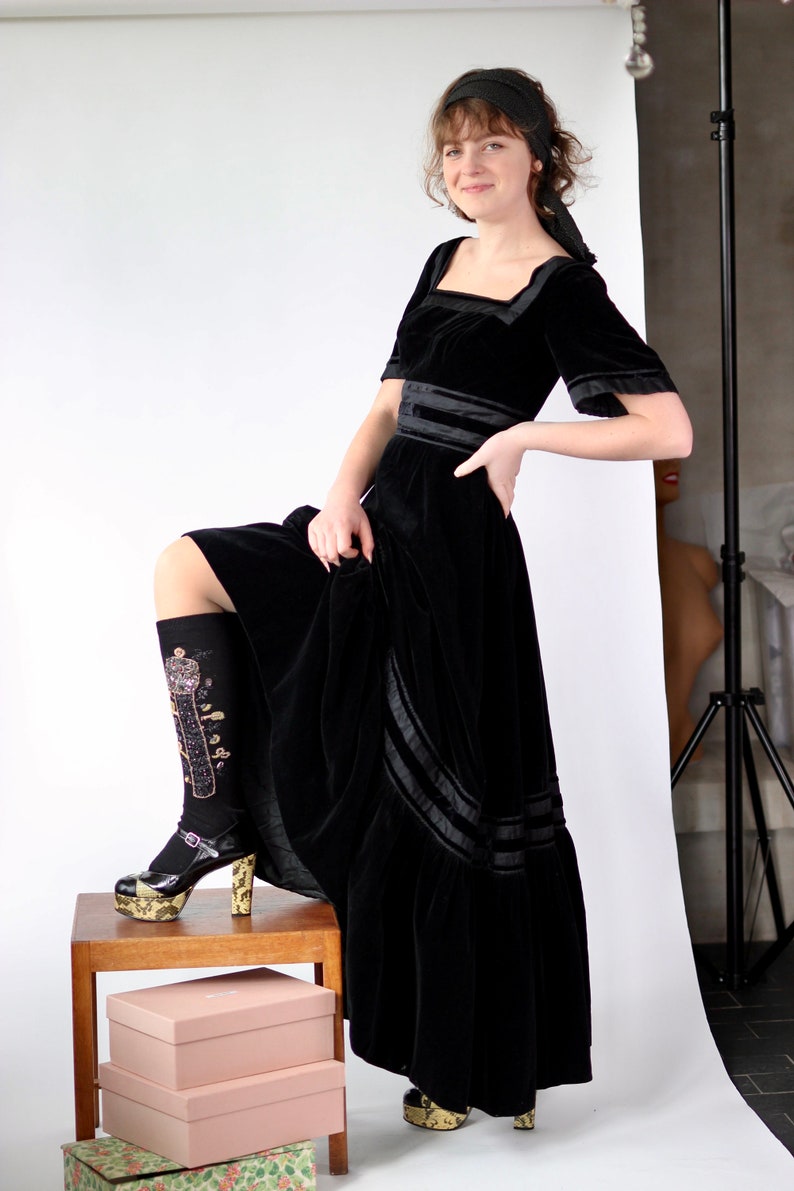 May include: A woman wearing a black velvet dress with a black velvet headband. She is standing on a wooden stool with two boxes stacked on top of each other. She is wearing black socks with gold embroidery and black platform shoes.