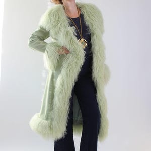 May include: A long, pale green coat with a fluffy, light green trim around the collar, cuffs, and hem. The coat is worn over a dark blue outfit and brown boots. A gold pendant necklace adds a touch of elegance.