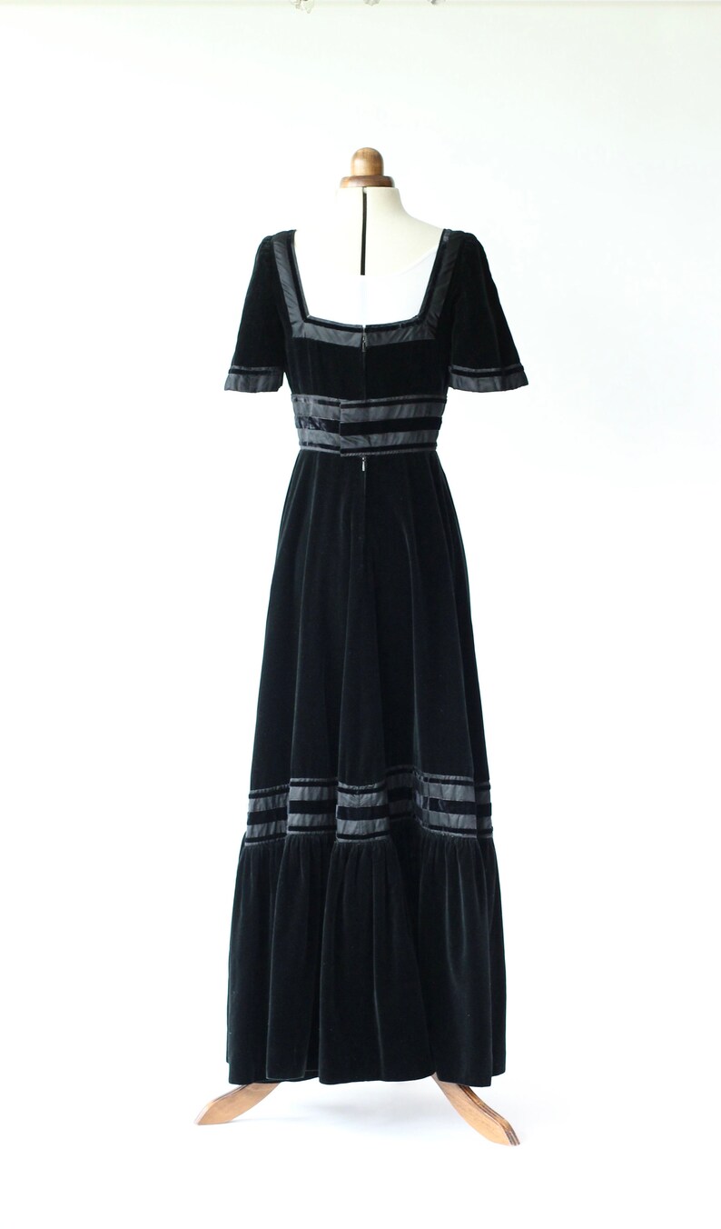 May include: A black velvet maxi dress with a square neckline and short sleeves. The dress features black satin ribbon trim around the neckline, waist, and hem.