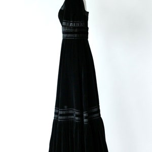 May include: A black velvet maxi dress with a square neckline and a fitted bodice. The skirt is full and flowing, with multiple layers of ruffles and black satin ribbon trim.