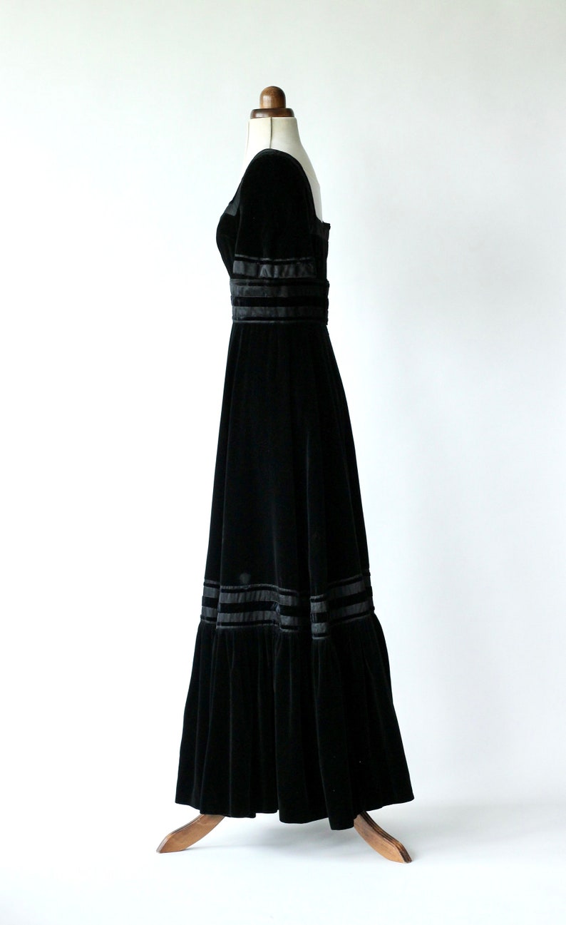 May include: A black velvet maxi dress with a square neckline and a fitted bodice. The skirt is full and flowing, with multiple layers of ruffles and black satin ribbon trim.