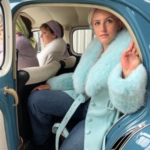 May include: Three women sitting in a vintage blue car. The woman in the back seat is wearing a light blue fur coat with a white fur collar. She is wearing blue jeans and has her hand raised. The woman in the front passenger seat is wearing a white jacket with a white fur collar and a pink and white headscarf. The woman in the driver's seat is wearing a purple jacket with a white fur collar and a green headscarf.