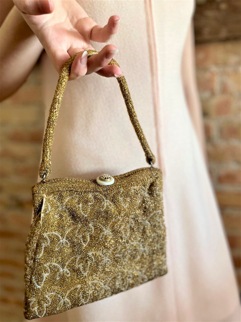 Puede incluir: Un bolso de noche de cuentas doradas con un cierre decorativo. El bolso tiene una correa corta y un dise&ntilde;o de cuentas.