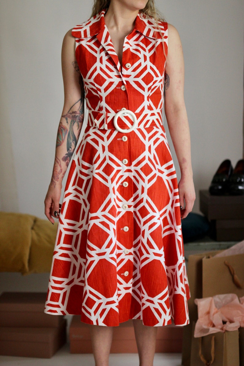 May include: A sleeveless orange and white geometric print dress with a white button closure and a white belt with a large oval buckle.