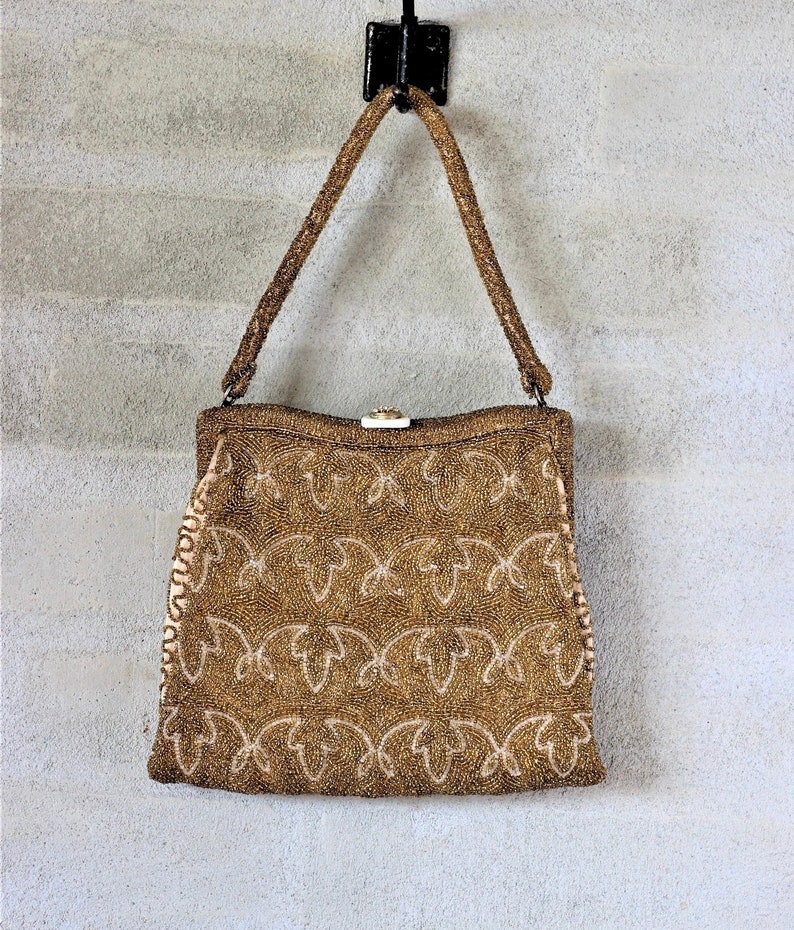 Puede incluir: Un bolso de mano de cuentas doradas con cierre de broche y un delicado patr&oacute;n floral. El bolso tiene una correa larga y est&aacute; colgado de un gancho.